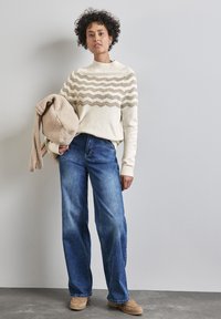 Cremfarbener Pullover mit gewellten beigen Streifen, kombiniert mit weit geschnittenen blauen Jeans. Beiger strukturierter Beutel wird am linken Arm gehalten, neutraler Hintergrund, sanfte Beleuchtung.