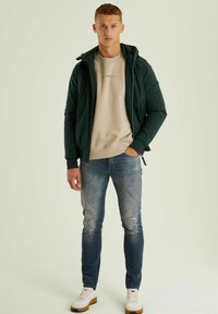 Temno zelen zip-up jakna čez bež kapuco, v kombinaciji z modrimi džinsom in belimi športnimi copati. Opazne teksture vključujejo gladko tkanino in džins.