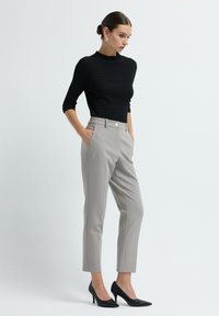 Woman wearing a fitted black textured top, light gray tailored ankle pants, black pointed high heels, and silver earrings, standing with hands in pockets.