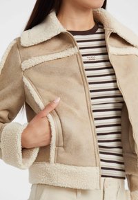 Giacca in finta suede beige con colletto e polsini di pelliccia crema, chiusura con zip e tasche laterali; indossata sopra una maglietta a righe marroni e bianche.