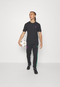 Camiseta negra con un pequeño logo morado, combinada con pantalones negros que tienen rayas verdes. El modelo sostiene un balón de fútbol multicolor y lleva zapatos negros.