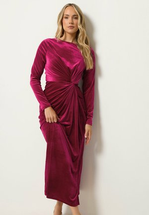 Mujer con cabello largo y rubio que lleva un vestido de terciopelo oscuro rosa con mangas largas y torcido, de pie contra una pared lisa y clara.
