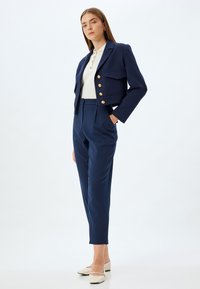 Marineblauwe cropped blazer met gouden knopen en twee voorzakken, gecombineerd met op maat gemaakte marineblauwe broek en crèmekleurige flats. Over het geheel een gladde textuur.