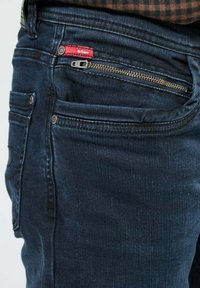 Jean bleu foncé avec une poche zippée horizontale à l'avant et une petite étiquette en tissu rouge près de la taille.