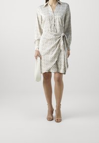 Femme portant une robe portefeuille beige à motifs avec des manches longues, tenant un sac à main blanc et portant des escarpins beige à bride de cheville.