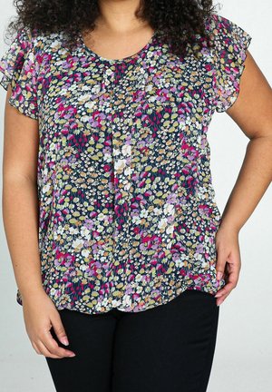 Femme portant un chemisier à manches courtes à motif floral multicolore et un pantalon noir, une main sur la hanche et l'autre détendue sur le côté.