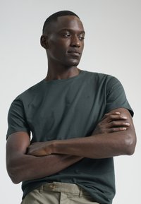 Camiseta corta de color verde oscuro, hecha de una tela suave, con cuello redondo y un pequeño logo en el pecho, combinada con pantalones color caqui claros.