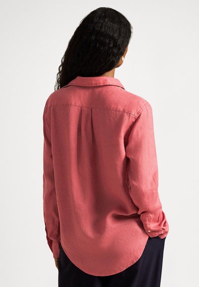 Sieviete ar gariem cirtainiem matiem, valkājot rozā krekliņu ar garām piedurknēm un pogām, redzama no muguras uz vienkrāsaina fona.