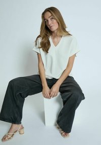 Camicia oversized bianca con scollatura a V abbinata a jeans ampi scuri e sandali con tacco nudo. Modella seduta su un piedistallo bianco.