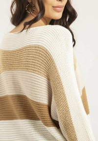 Pull en tricot avec des rayures horizontales en crème et beige doré. Coupe ample avec un col large et un tissu texturé. Accessoires visibles.
