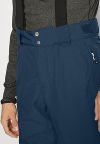 Blaue wasserdichte Hosen mit verstellbaren Trägern, Metallknöpfen und einer mit Reißverschluss versehenen Tasche. Glatte Textur mit geradem Schnitt.