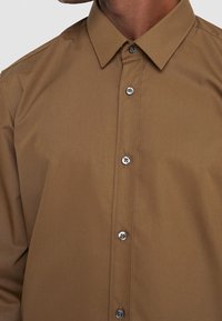 Hnědá košile na knoflíky s klasickým límcem, hladkou texturou a stříbrnými knoflíky vpředu.