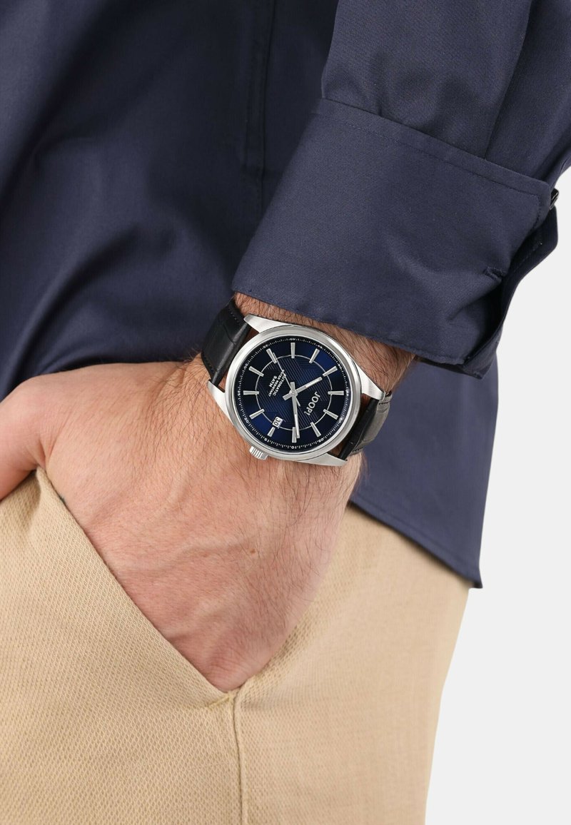 Silberne Uhr mit einem dunkelblauen Zifferblatt, schwarzem Lederarmband, Datumsanzeige und silbernen Stundenmarkierungen, getragen an einem Handgelenk mit einem marineblauen Hemd.