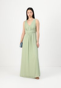 Hosszú, lebegő zöld ruha V-nyakkal, redőzött derékkal és puha anyaggal. Kék clutch táskával és lapos szandálokkal kísérve.