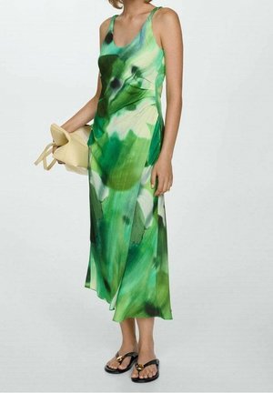 Femme portant une robe midi sans manches verte et blanche, tenant une pochette beige et portant des sandales noires avec des détails dorés.