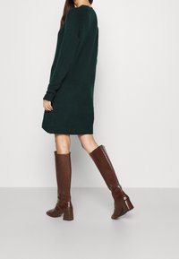 Robe tricotée vert foncé avec de longues manches et un col arrondi, associée à des bottes hautes en cuir verni marron à imprimé crocodile qui montent jusqu'au genou.