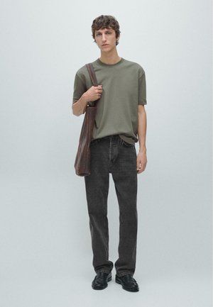 Jeune homme portant un t-shirt vert olive, jean noir, chaussures noires, tenant un sac à bandoulière marron sur un fond clair uni.