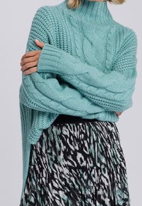 Pull turquoise clair en maille torsadée avec un col montant, porté sur une jupe plissée noire à motifs teal, avec des manches larges et oversize.