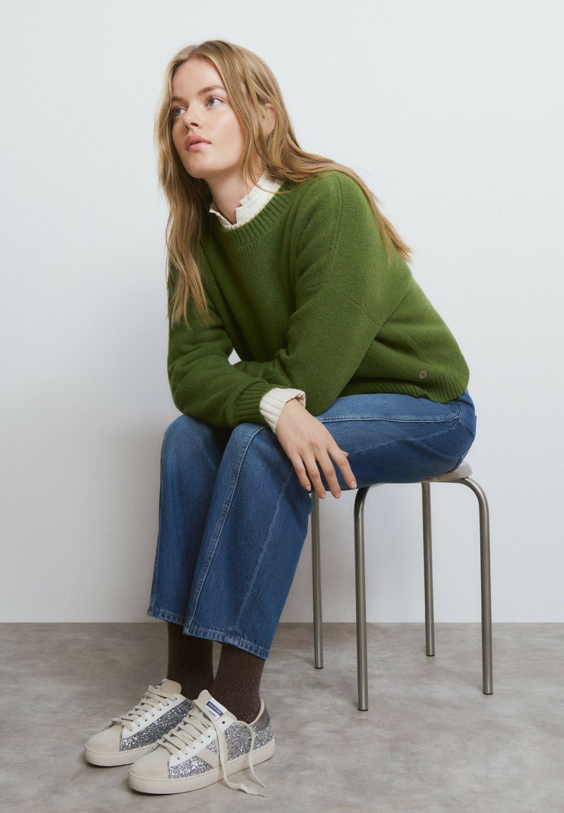Jersey de punto verde con cuello blanco, vaqueros azul de pierna ancha y zapatillas beige con detalles plateados, sentada en una silla metálica.