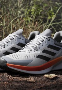 Baskets de sport gris clair avec des rayures noires et des accents orange. Tige texturée avec des trous de ventilation et un design de semelle robuste.