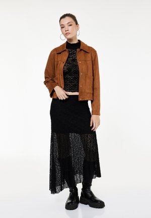 Mujer con una chaqueta marrón sobre un top corto de encaje negro y falda, con botas negras gruesas, posando sobre un fondo blanco.