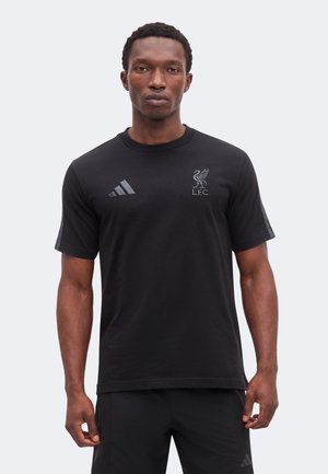 Homme portant un t-shirt noir Liverpool FC Adidas et un short noir, debout avec une expression neutre sur un fond uni.