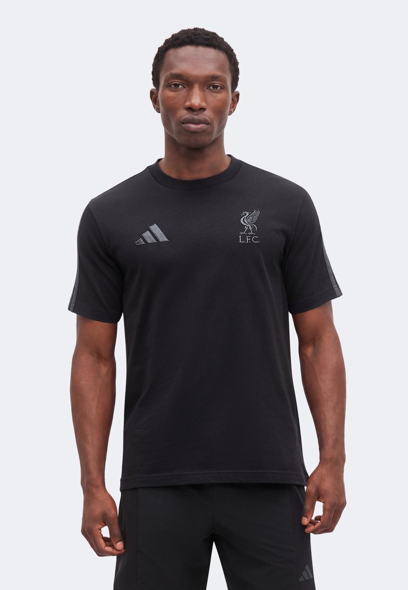 Άνδρας που φοράει μαύρο t-shirt Liverpool FC Adidas και μαύρο σορτς, στέκεται με ουδέτερη έκφραση μπροστά σε απλό φόντο.