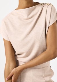 Ljust rosa draperad topp med mjuk ribbad textur, med en båt- halsringning och tre dekorativa knappar på axeln.