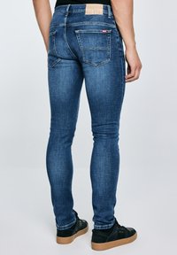 Egy férfi, aki vékony szabású kék farmert és fekete tornacipőt visel, sima világos háttér előtt áll, hátsó nézetben.