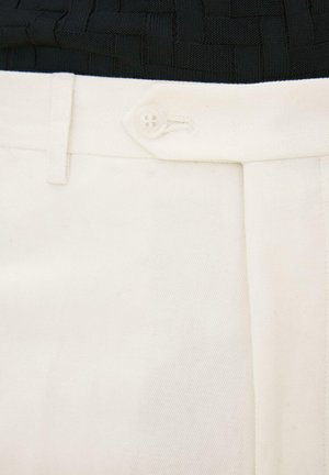 Nærbillede af taljebåndet på hvide bukser med knaplukning og bæltestrop, kombineret med en sort tekstureret top.