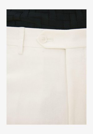 Nahaufnahme des Taillenbunds weißer Hosen mit Knopfverschluss und Gürtelöse, kombiniert mit einem schwarzen strukturierten Oberteil.