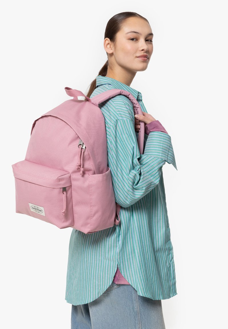 Mochila rosa con una superficie texturizada, forma redondeada, bolsillo frontal y correas ajustables, llevada por una persona con una camiseta a rayas.