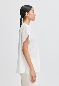 Haut blanc à manches courtes avec une coupe décontractée, présentant un tissu texturé et des fentes sur les côtés. Associé à un pantalon de couleur claire.