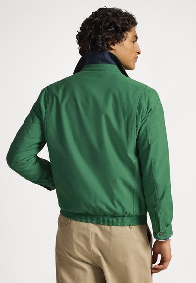 Πράσινο μπουφάν μπόμπερ με σκούρο μπλε γιακά, ελαστική μέση και μανσέτες με κουμπιά. Λείο ύφασμα και απλός σχεδιασμός. Φορεμένο πάνω από χακί παντελόνια.