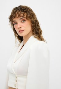 Blazer blanco con escote en V profundo y cierre de botón; presenta mangas anchas y fluidas y una textura de tela suave.