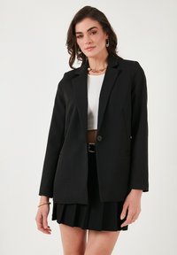 Crni sako s reverima i jednim gumbom, uparen s rebrastim bijelim topom i plisiranim crnim suknjom. Glatka tekstura tkanine.