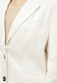 Hellbeige Blazer aus strukturiertem Stoff mit Fischgrätenmuster, ausgestattet mit einer einzelnen Brusttasche und schwarzen Knöpfen.