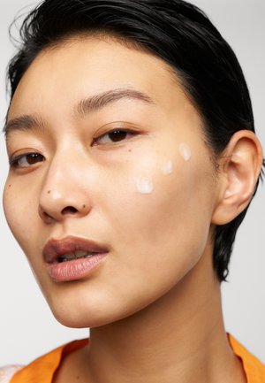 Primo piano di un volto con pelle liscia, caratterizzato da tre piccole gocce di crema sulla guancia e una leggera lucentezza. I capelli sono corti e scuri.