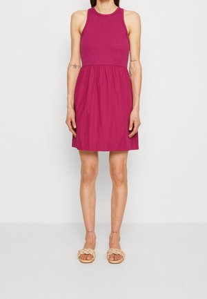 Kobieta w różowej, bezrękawnikowej sukience do kolan i beżowych plecionych sandałach, stojąca na tle białej ściany.