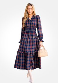 Largo vestido de cuadros con colores azul marino oscuro, rojo y verde; cintura fruncida; dobladillo con volantes; combinado con un bolso beige y botas de color claro.