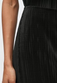 Το ύφασμα του μαύρου πλισέ φορέματος παρουσιάζει κατακόρυφες ρίγες, λεία υφή και ελαφρώς συγκρατημένη ζώνη στη μέση με κρυφό πλαινό ραφάκι.