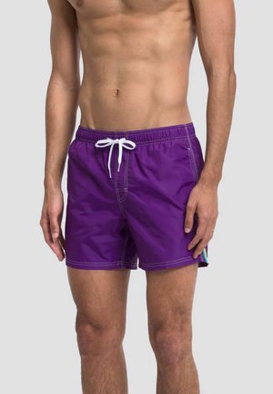 Torso e gambe maschili che indossano pantaloncini da bagno viola con coulisse bianca e cuciture visibili, in piedi su uno sfondo chiaro e uniforme.