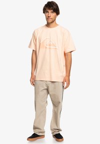 Lys ferskenfarget bomulls T-skjorte med en hvit grafisk logo, kombinert med lyse beige cordfløyelsbukser og svarte slip-on sko.