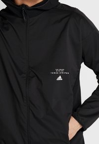 Svart zip-up jacka i slät, lättviktig tyg med huva, diskreta meshinslag och vit branding på bröstet.