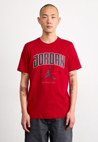 Crvena majica kratkih rukava s natpisom "JORDAN" ispisanim podebljanim sivim slovima, s siluetom figure koja zakucava u crnoj boji. Pamuk.