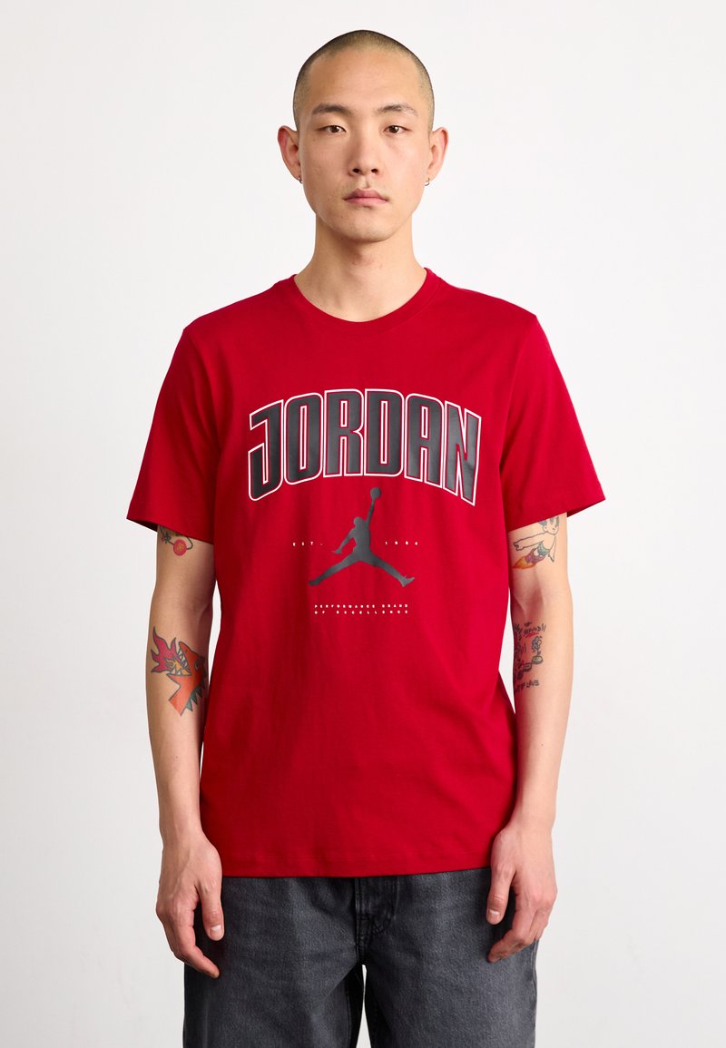 Crvena majica kratkih rukava s natpisom "JORDAN" ispisanim podebljanim sivim slovima, s siluetom figure koja zakucava u crnoj boji. Pamuk.