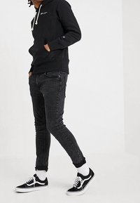 Sudadera negra con capucha y cordones, jeans ajustados grises y zapatillas negras. Calcetines blancos son visibles; atuendo casual con un ajuste relajado.