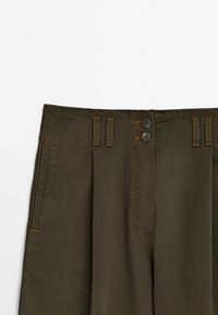 Pantalons vert olive foncé avec plis, deux poches avant, passants de ceinture et fermeture à double bouton avec couture contrastante.