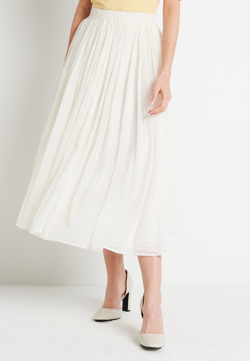 Femme portant une jupe midi plissée blanche, un haut beige et des chaussures à talons hauts beige clair, debout sur un sol blanc contre un mur blanc.