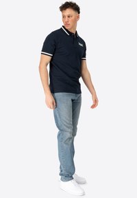 Mørkeblå polo-shirt med hvide striber på kraven og ærmerne, parret med lyseblå jeans og hvide sneakers, på en neutral baggrund.